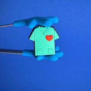 Może przedstawiać: Turkusowy, miniaturowy urok chirurga z czerwonym sercem i dwiema kieszeniami. Urok jest trzymany przez niebieski instrument medyczny na niebieskim tle. Fartuch ma dekolt w kształcie litery V.