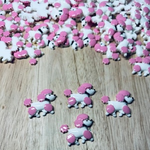 Peut inclure: Un tas de breloques en forme de caniche blanches et roses avec des yeux noirs. Les breloques sont éparpillées sur une surface en bois. Il y a quatre breloques au premier plan, avec une fourrure rose et un corps blanc.