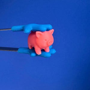 Puede incluir: Una pequeña figura de cerdo rosa es sostenida por pinzas azules y plateadas sobre un fondo azul. El cerdo tiene detalles visibles, incluyendo orejas, hocico y patas. Las pinzas tienen un diseño único en forma de mano.
