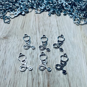 Może przedstawiać: Sześć srebrnych charmów w kształcie stetoskopu na drewnianej powierzchni. Charms są małe i szczegółowe, z realistycznym wzorem stetoskopu.