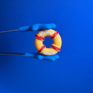 Peut inclure: Un bouée de sauvetage jaune avec des détails de corde rouge et les mots "WELCOME ABOARD" en lettres blanches. La bouée est tenue par des pinces bleues sur un fond bleu.