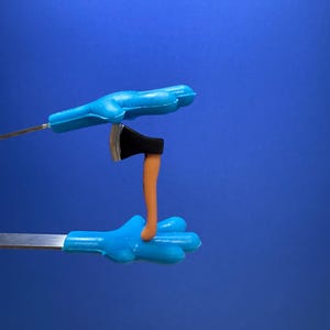 May include: A miniature toy axe with a black blade and orange handle is held by two blue plastic hands on metal rods against a blue background. The axe is positioned between the hands.