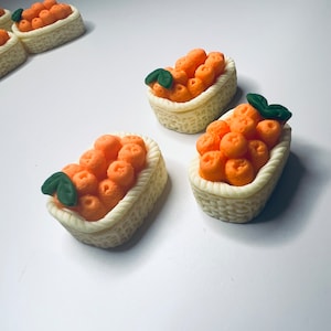 May include: Four miniature orange baskets filled with orange fruit, each with a green leaf on top. The baskets are made of white clay and have a woven texture.