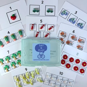 May include: Educational flashcards with illustrations and small objects. The cards show numbers 1-10 with images of an apple, trucks, bicycles, cacti, pencils, tanks, trees, flowers, bananas, and sheep. A clear plastic box is in the center. The website speechandsmile.com is visible.