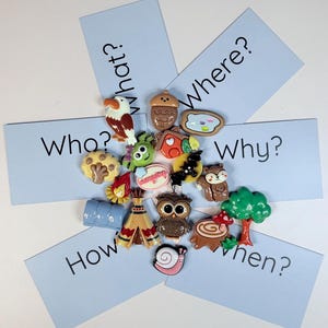 Peut inclure: Une collection d'épingles et de breloques colorées et fantaisistes, dont un hibou, un arbre et un escargot, disposées autour de cartes bleu clair avec des mots interrogatifs comme "Qui ?" et "Quoi ?".