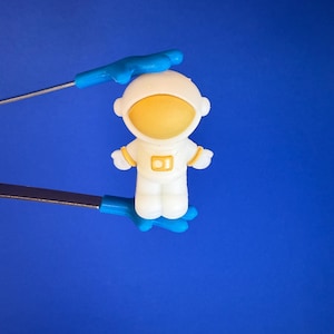 Peut inclure: Figurine d'astronaute blanc avec un visage et des détails jaunes, tenue par des pinces bleues et argentées sur fond bleu. L'astronaute a un casque et une combinaison blancs avec une visière jaune et la lettre "B" sur la poitrine.