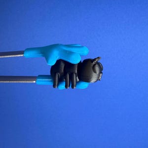 May include: A black ant-shaped object is held by blue tongs against a blue background. The tongs have silver metal handles and blue plastic hand grips. The ant has a rounded body and detailed legs and antennae.