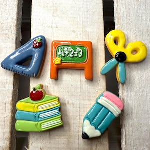 Peut inclure: Ensemble d'aimants colorés sur le thème de l'école. Comprend un triangle bleu avec une fraise, un tableau orange avec des chiffres, des ciseaux jaunes, une pile de livres avec une pomme et un crayon bleu sarcelle avec une gomme rose.