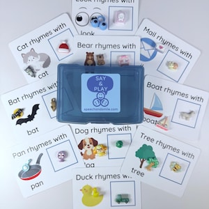 Puede incluir: Juego de tarjetas educativas de rimas con ilustraciones y objetos en miniatura. Las tarjetas presentan palabras como "gato", "murciélago" y "pato", con imágenes correspondientes y pequeños adornos coloridos. Incluye una caja de almacenamiento azul.
