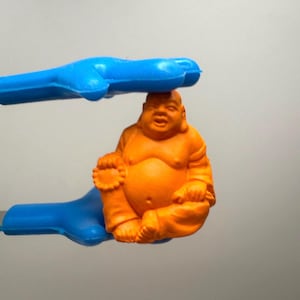 Puede incluir: Una figura de Buda naranja es sostenida por unas pinzas de plástico azules. La figura está en posición sentada con una expresión sonriente. Las pinzas son de color azul brillante.