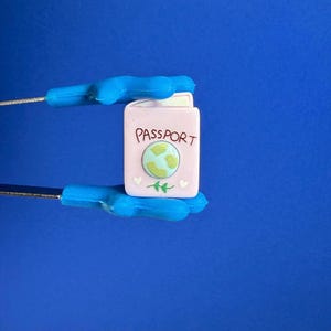 Peut inclure: Un charm en forme de passeport rose clair avec le mot "PASSPORT" imprimé sur le devant. Le charm présente un globe vert et bleu, deux brins verts et deux cœurs blancs. Tenue par des pinces bleues sur fond bleu.