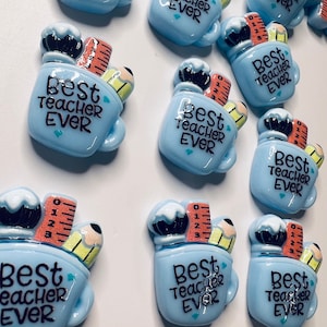 Può includere: Una collezione di 10 ornamenti in ceramica a forma di tazza, di colore blu e bianco, con una matita, un righello e una gomma da cancellare all'interno. Ogni tazza ha il testo "Best Teacher Ever" sul davanti.