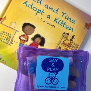 May include: A purple plastic container with a blue sticker that says "SAY & PLAY" with three smiley faces and the website speechandsmile.com. The container has a variety of items inside, including a blue ribbon, a white card, and a pink bottle. The container is sitting on top of a yellow book titled "Ted and Tina Adopt a Kitten" with illustrations of children and cats.