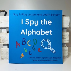 May include: A blue book with the title "I Spy the Alphabet" and a magnifying glass with colorful letters scattered around it. The book is part of the "Say & Play Letters and Learn Series".