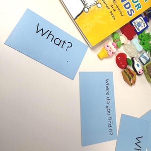 May include: A collection of educational materials, including a children's book and blue flashcards. The flashcards have the questions "What?" and "Where do you find it?" printed on them. Various miniature food and object toys are scattered around the book.