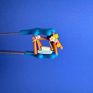 Peut inclure: Une balançoire miniature avec un siège bleu clair et un cadre marron, ornée de fleurs colorées. La balançoire est tenue par des pinces en métal à poignées bleues sur un fond bleu uni. C'est un objet décoratif.