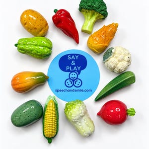 May include: A collection of twelve miniature plastic vegetable figurines arranged in a circle around a blue circular logo that reads SAY & PLAY speechandsmile.com. The set includes a potato, red pepper, broccoli, corn, cauliflower, okra, radish, cabbage, and squash.