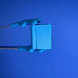May include: A light blue, 3D-printed cube held by two blue plastic handles with metal rods. The cube has vertical lines on its surface. The background is a solid blue.