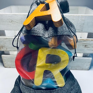 May include: A grey burlap drawstring bag filled with colourful wooden letter blocks. The bag is tied with a black cord and has a clear mesh lining. The letters are visible through the mesh and include the letters A, C, and R.