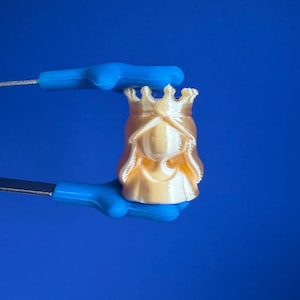 May include: A small, gold-colored figurine of a queen wearing a crown. The figurine is held by a blue tool against a solid blue background. The queen's face is smooth, and the crown has intricate details.