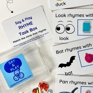 May include: A Say & Play RHYME Task Box set with a clear plastic container, white cards featuring rhyming words like duck, look, bat, and pan with corresponding illustrations, and a collection of small colorful plastic miniature objects.