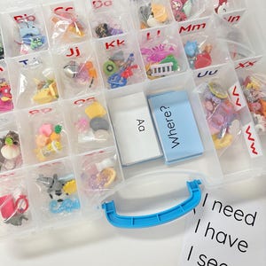 May include: A clear plastic storage organizer with a blue handle, featuring multiple compartments labeled with alphabet letters. Each section contains small plastic trinkets in individual bags. Two white cards are visible with the text Aa and Where?