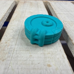 Puede incluir: Un objeto tridimensional circular de color verde azulado que presenta un borde elevado con detalles segmentados y una pequeña hendidura cuadrada en la superficie superior. El objeto tiene una estructura escalonada tipo escalera.