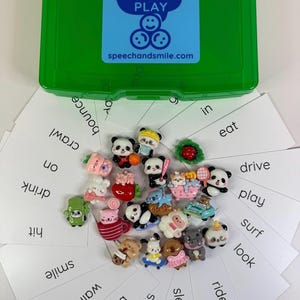 May include: A green plastic box with the text "SAY & PLAY" and "speechandsmile.com" on a blue sticker. The box is surrounded by white cards with action words and small animal figurines.