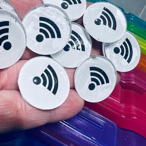 Könnte beinhalten: Eine Hand hält eine Gruppe weißer runder Aufkleber mit einem schwarzen WLAN-Symbol auf jedem. Die Aufkleber befinden sich in einem durchsichtigen Plastikbehälter.