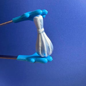 May include: A small, white, 3D-printed bowling pin held by a pair of bright blue, cartoon-style hand-shaped tongs. The bowling pin features a smooth, pearlescent finish with visible vertical layer lines from the printing process, set against a solid blue background.