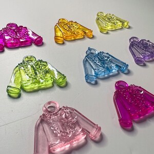 May include: A collection of colorful, translucent jacket-shaped charms in various shades including pink, yellow, green, and blue. Each charm features a detailed floral design on the front and a small loop at the top for hanging.