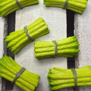 Może przedstawiać: Sześć pęczków zielonych szparagów związanych brązowymi gumkami na białym drewnianym tle.