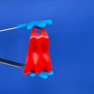May include: A miniature red cape with a blue handle, designed for a toy or figurine. The cape is a vibrant red, and the handle is a bright blue. The background is a solid blue.