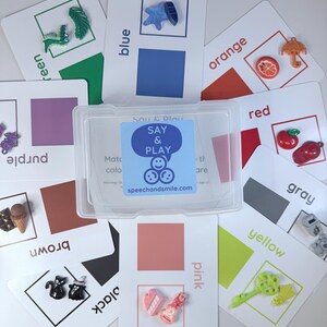 Könnte beinhalten: Eine Sammlung von Farbkarten mit kleinen, farbigen Objekten. Jede Karte zeigt einen Farbnamen und ein entsprechendes Quadrat. Das Set enthält eine transparente Kunststoffbox mit dem Text "SAY & PLAY" und der Website speechandsmile.com.