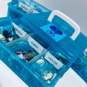 May include: A clear blue plastic storage box with a white handle and multiple compartments. Each compartment is labeled with a white label. The labels read "FOREST", "ANIMALS", "FARM", "HOUSE", and "PRE". The box has a sticker that reads "SAY & PLAY speechandsmile.com".