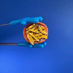 May include: A miniature round orange basket filled with yellow french fries, held by blue plastic tongs. The fries have small green specks, and the basket features a textured, woven-look rim. The entire object is presented against a solid blue background.