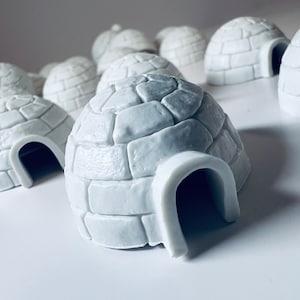 Puede incluir: Una colección de pequeños objetos decorativos blancos con forma de iglú. Cada iglú tiene una cúpula redondeada con un patrón texturizado similar a ladrillos y una entrada curva. Los objetos están dispuestos sobre una superficie blanca.