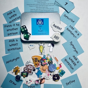 Peut inclure: Un jeu "Say & Play" pour adolescents, comprenant une boîte en métal, des cartes de mots, des breloques et des dés. L'ensemble comprend des cartes avec des invites comme "Posez une question QUAND" et des breloques comme une part de pizza et un vélo.