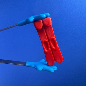May include: A pair of kitchen tongs featuring bright blue handles shaped like cartoon hands with four fingers. The metal arms hold two vertical red plastic objects that have a smooth, flat surface with a small oval indentation in the center.