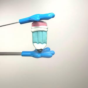 Peut inclure: Une gomme fantaisiste en forme de crayon, avec un corps bleu clair, un dessus de gomme rose et blanc, et une pointe blanche avec une pointe noire. La gomme est tenue par deux supports bleus en forme de main avec des tiges métalliques.