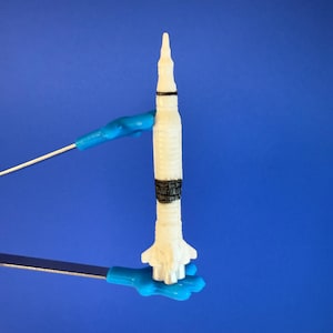 Peut inclure: Une maquette de fusée blanche avec des bandes noires et grises, maintenue par des supports bleus sur un fond bleu. La fusée a un sommet conique et un corps cylindrique détaillé, sur le thème de l'exploration spatiale miniature.