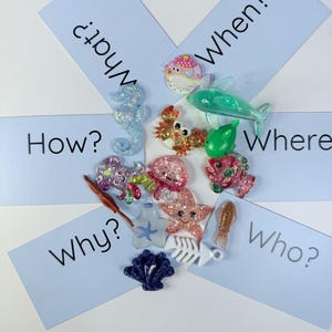 Puede incluir: Un surtido de adornos brillantes con forma de criaturas marinas, incluyendo un caballito de mar, un cangrejo, una estrella de mar y un pez, se disponen alrededor de tarjetas azul claro con palabras interrogativas.