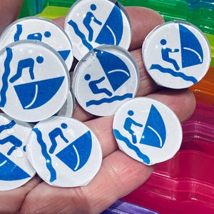 Puede incluir: Una colección de azulejos redondos de vidrio blanco con ilustraciones azules y blancas de una persona navegando en un velero. Los azulejos están dispuestos en una mano.