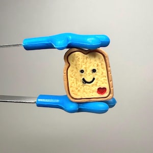 May include: A slice of toast with a smiling face and a red heart, held by blue tongs. The toast has a light brown crust and a yellow center. The tongs are made of metal and blue plastic.