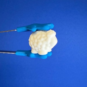 May include: A white sheep-shaped object with a textured surface, held by blue tongs against a blue background. The sheep has a small patch of blue on its back. The tongs have blue handles and silver metal prongs.