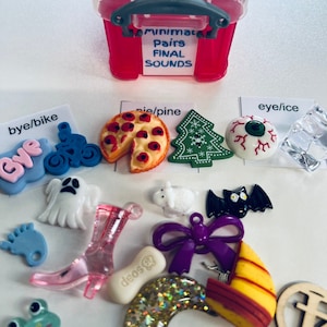 Op de afbeelding: Een roze plastic doos met de woorden "SAY & PLAY" en "Minimal Pairs FINAL SOUNDS" erop. De doos is gevuld met verschillende kleurrijke speelgoed, waaronder een spook, een fiets, een pizza, een kerstboom, een oog, een kikker, een schoen, een boot, een strik, een vleermuis, een hondenbot, een halve maan en een kruis.