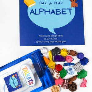 May include: A blue book titled Say & Play Alphabet by Jill Bacigalupi, Speech-Language Pathologist, alongside a blue plastic pouch labelled Talkets by Speech & Smile containing various small, colourful, shaped objects including a key and animals.