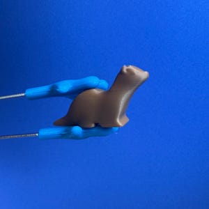 Puede incluir: Una figura de hurón marrón impresa en 3D sostenida por pinzas metálicas con mangos azules sobre un fondo azul. El hurón está en una pose de pie, con el cuerpo ligeramente curvado. Las pinzas sujetan el hurón por los lados.