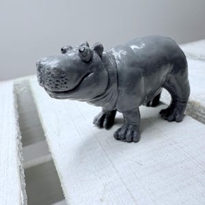 Peut inclure: Figurine d'hippopotame grise souriante. L'hippopotame est debout sur ses quatre pattes, avec des pieds détaillés et une surface texturée. La figurine est de couleur unie et semble être faite d'un matériau durable.