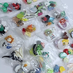 Puede incluir: Surtido de pequeños amuletos de plástico de colores en bolsas de plástico transparentes individuales. Los amuletos presentan diversos diseños, incluyendo animales, comida y objetos.